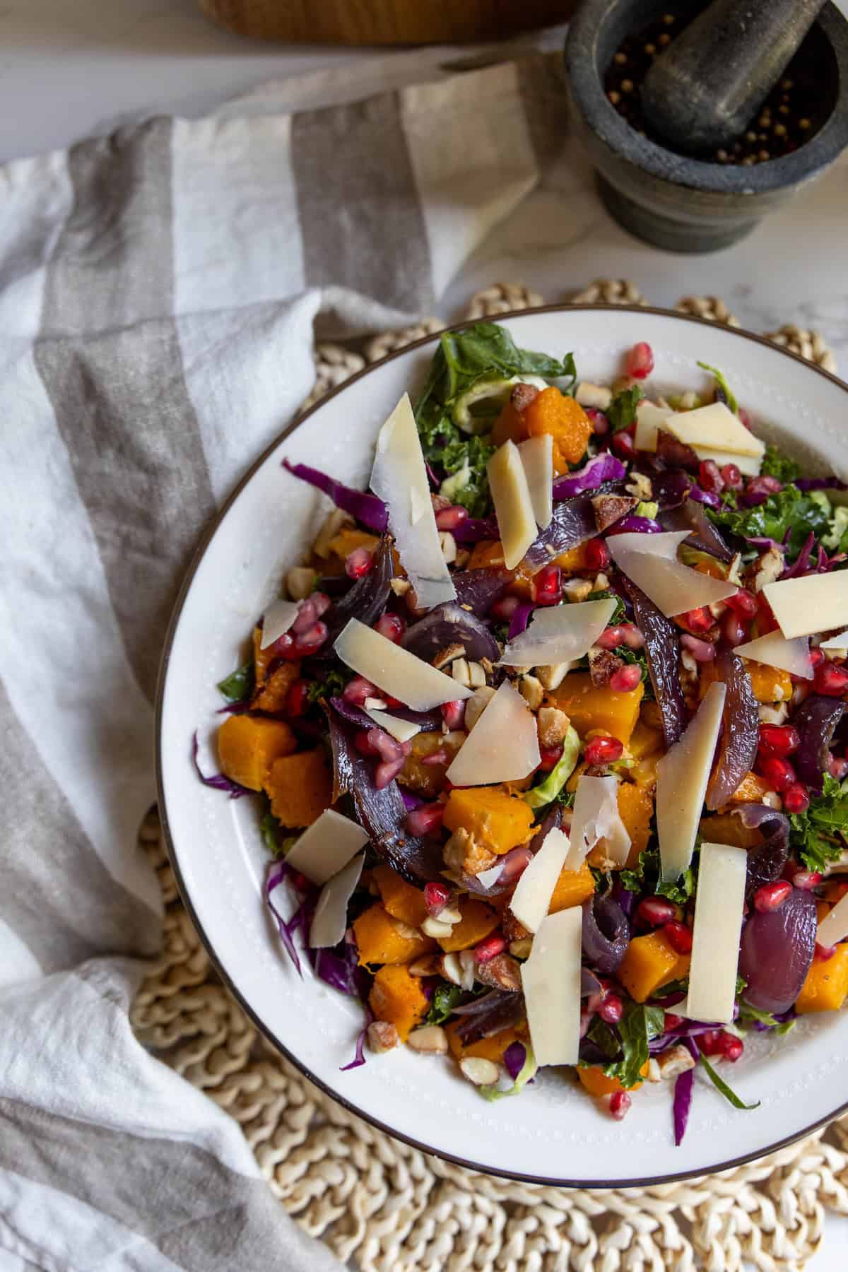 A plate of winter salad with mixed greens, roasted butternut squash, red onions, pomegranate seeds, and shaved cheese sits on a woven placemat.