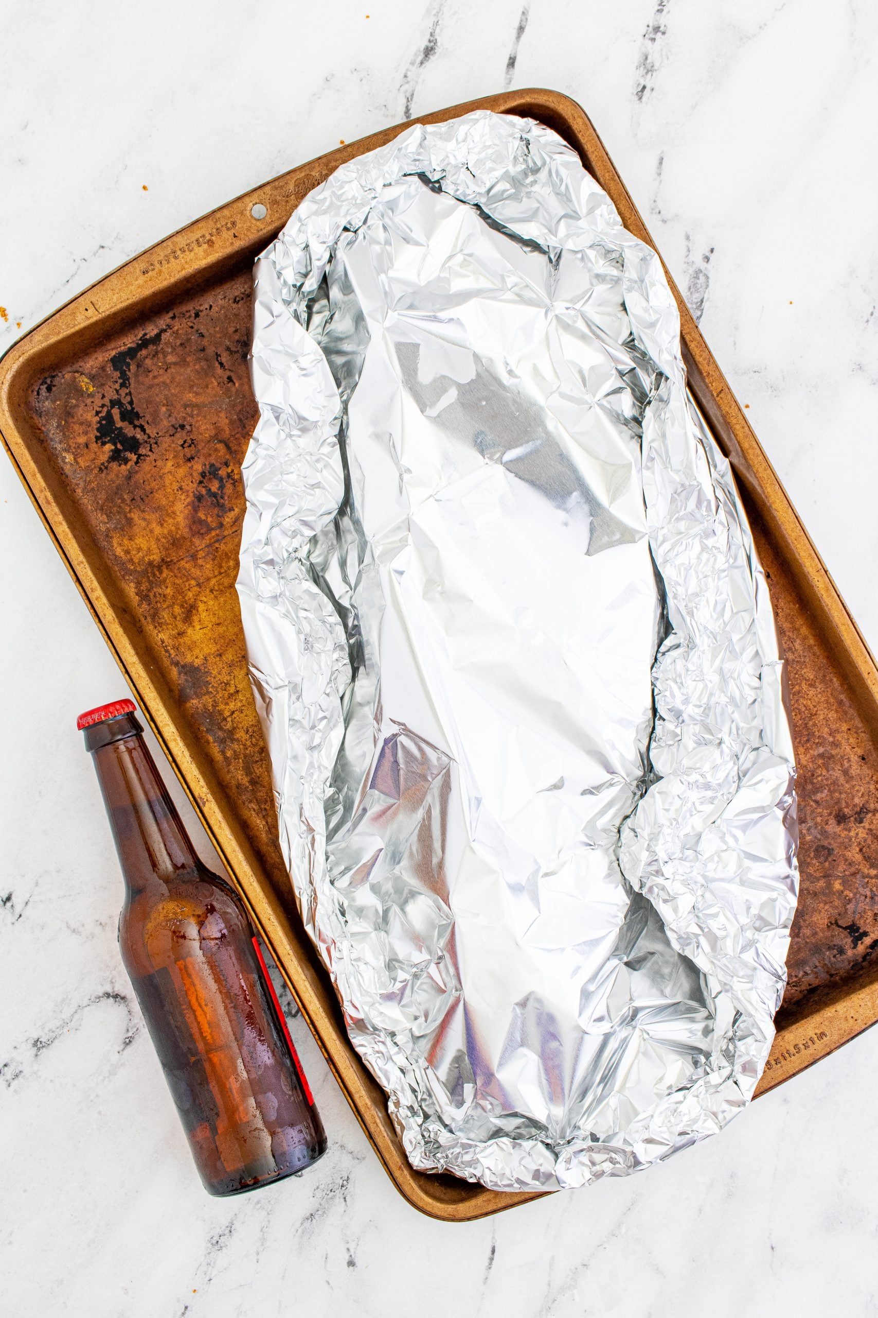 ribs wrapped in aluminum foil on a baking sheet with a bottle of beer sitting next to it
