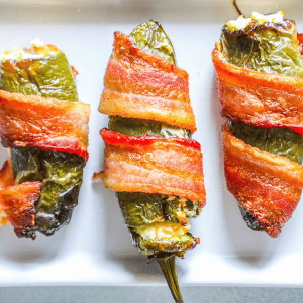 Three bacon-wrapped cheddar and garlic jalapeรฑo poppers filled with cheese sit on a white plate, offering an easy low-carb treat perfect for keto enthusiasts.