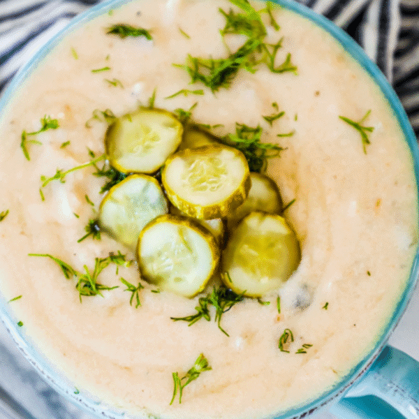 A close-up of a creamy pickle soup garnished with sliced pickles and chopped fresh herbs in a blue bowlโa delicious twist on the classic dill pickle soup recipe.