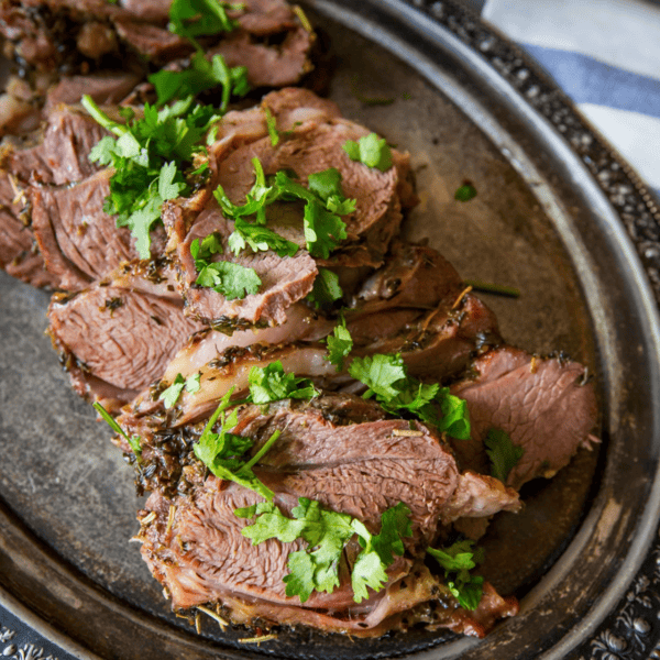 Sliced roasted leg of lamb, garnished with herbs, rests elegantly on an oval metal platter, showcasing the best easy leg of lamb preparation.