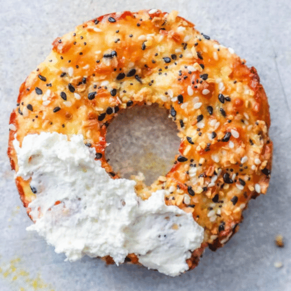 A partially eaten Keto Everything Bagel with a thick layer of cream cheese rests on a light gray surface.