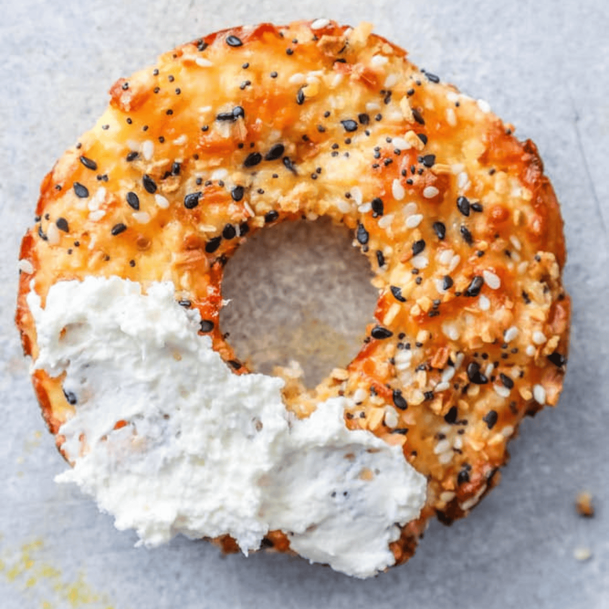 A partially eaten Keto Everything Bagel with a thick layer of cream cheese rests on a light gray surface.