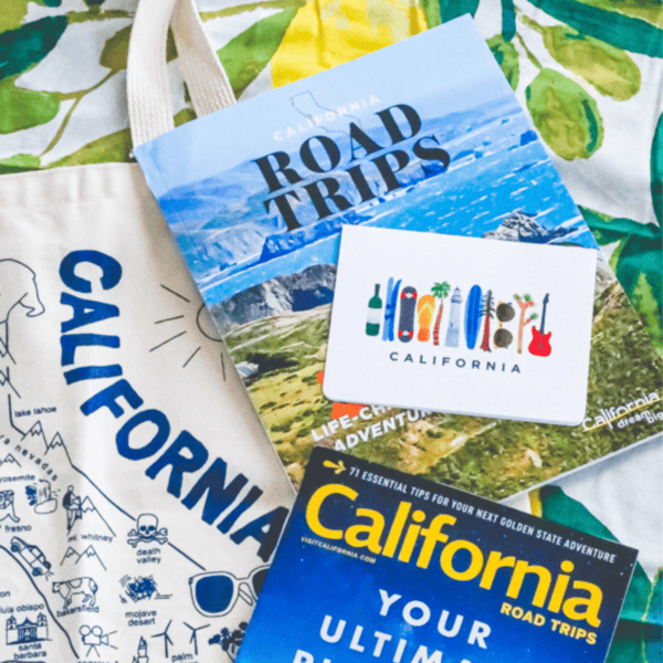 A collection of California-themed travel items, perfect for anyone looking to Road Trip California, includes a tote bag, two guidebooks titled "California Road Trips," and a postcard displaying various colorful illustrations.